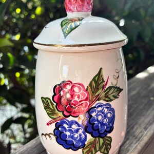 May include: A cream-colored ceramic jar with a lid, adorned with hand-painted raspberries and leaves. The lid features a raspberry-shaped knob. Gold trim accents the rim and lid. The jar is approximately 5 inches tall.