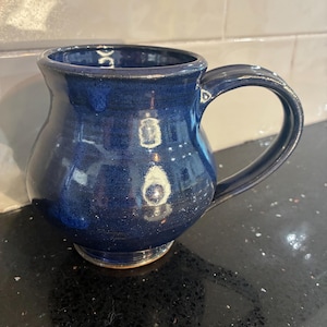 May include: A handmade, navy blue ceramic mug with a rounded body and a large, curved handle. The mug has a glossy finish and a slightly flared rim. The base of the mug has a lighter, tan color.