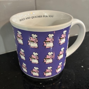 Può includere: Tazza in ceramica bianca con una fascia viola con maiali dei cartoni animati con cappelli da chef che tengono quiche. La tazza ha il testo "HOGS AND QUICHES FOR YOU" stampato sul bordo interno. La tazza ha un manico bianco.
