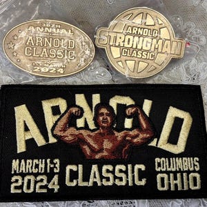 May include: Two gold-colored metal pins and a black patch with gold lettering and a muscular man graphic. The pins read "Arnold Strongman Classic" and "36th Annual Arnold Classic 2024." The patch says "Arnold Classic Columbus Ohio March 1-3 2024."