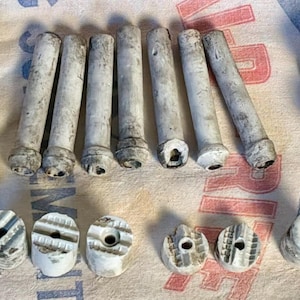 May include: A collection of vintage ceramic electrical insulators and pins. The insulators are white with a cylindrical shape and a central hole. The pins are long, cylindrical, and have a weathered appearance. Some insulators have the text "WF 36".