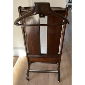 May include: A dark brown wooden valet stand with a built-in hanger, a small shelf, and a lower bar. The stand has a rectangular shape and is mounted on small wheels for easy movement. The wood grain is visible.