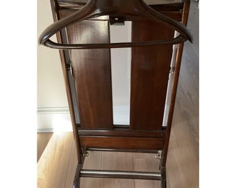 May include: A dark brown wooden valet stand with a built-in hanger, a small shelf, and a lower bar. The stand has a rectangular shape and is mounted on small wheels for easy movement. The wood grain is visible.