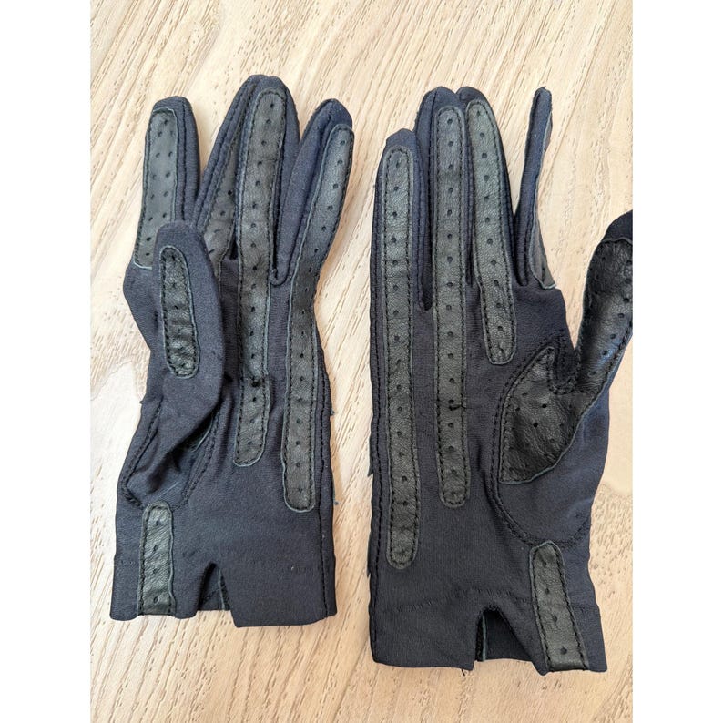 May include: A pair of black gloves with perforated leather detailing. The gloves feature a classic design with finger and palm accents. The gloves are made of a soft, dark material, suitable for driving or fashion.