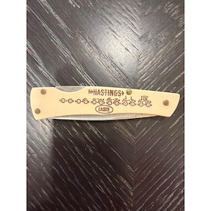 May include: A cream-colored pocket knife with a stainless steel blade. The knife features the word "HASTINGS" and "CASITE" etched on the handle, along with a train design. The knife is secured with brass rivets.