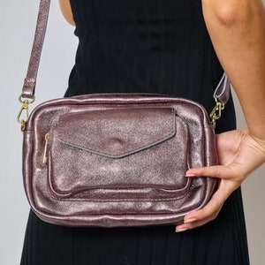 Puede incluir: Un bolso de hombro rosa metálico con un bolsillo con cremallera en la parte delantera. El bolso tiene una correa de cadena dorada y un tirador de cremallera dorado.