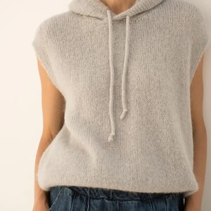 May include: A light grey sleeveless hoodie with a drawstring. The hoodie is made of a soft, textured knit material. The model is wearing blue denim trousers. The overall style is casual and comfortable.