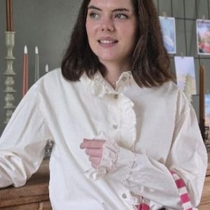 以下が含まれることがあります： フリルカラーと袖口のクリーム色のブラウス。長袖シャツはボタン留めで、濃い青色のスカートと組み合わせています。ピンクと白のストライプのバッグストラップが見えます。