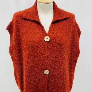 May include: A sleeveless, rust-coloured knitted vest with a collared neckline and three large, round, wooden buttons. The vest is displayed on a mannequin and has a soft, textured appearance. The collar is a darker shade of rust.