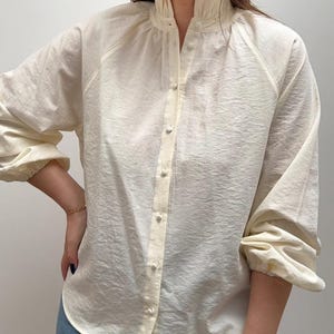 May include: A cream-colored long-sleeve button-down shirt featuring a high ruffled collar, a front placket with small matching buttons, and gathered cuffs. The fabric has a subtle textured finish and a relaxed fit with raglan-style sleeves.