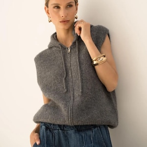 Pode incluir: Um suéter cinza sem mangas com capuz e fecho de zíper completo. Combinado com calças jeans azuis. A pessoa está usando brincos de argola dourados e uma pulseira dourada. O conjunto é casual e confortável.
