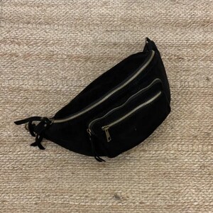 Peut inclure: Sac banane noir avec deux compartiments zippés et une petite poche avant. Le sac a une texture semblable à du daim et des fermetures éclair dorées. La sangle est réglable et possède une fermeture à boucle.