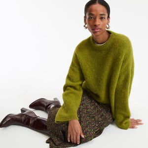 May include: A woman wearing a green sweater, floral skirt, and brown boots. The sweater is a solid, textured green. The skirt has a dark background with a floral pattern. The boots are a dark brown color.
