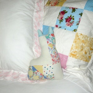 May include: A patchwork quilt with a floral design in shades of blue, pink, yellow, and white. A patchwork toy in the shape of a bunny is lying on the quilt.