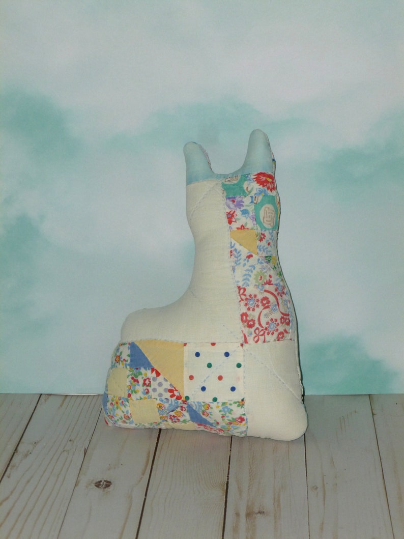 May include: A white patchwork fabric llama toy with a blue, yellow, red, and green floral pattern. The toy is sitting on a light wood surface.