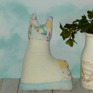 May include: A handmade patchwork fabric toy in the shape of a llama. The llama is light blue with yellow, pink, and white patchwork fabric. The llama is sitting on a light wood surface.