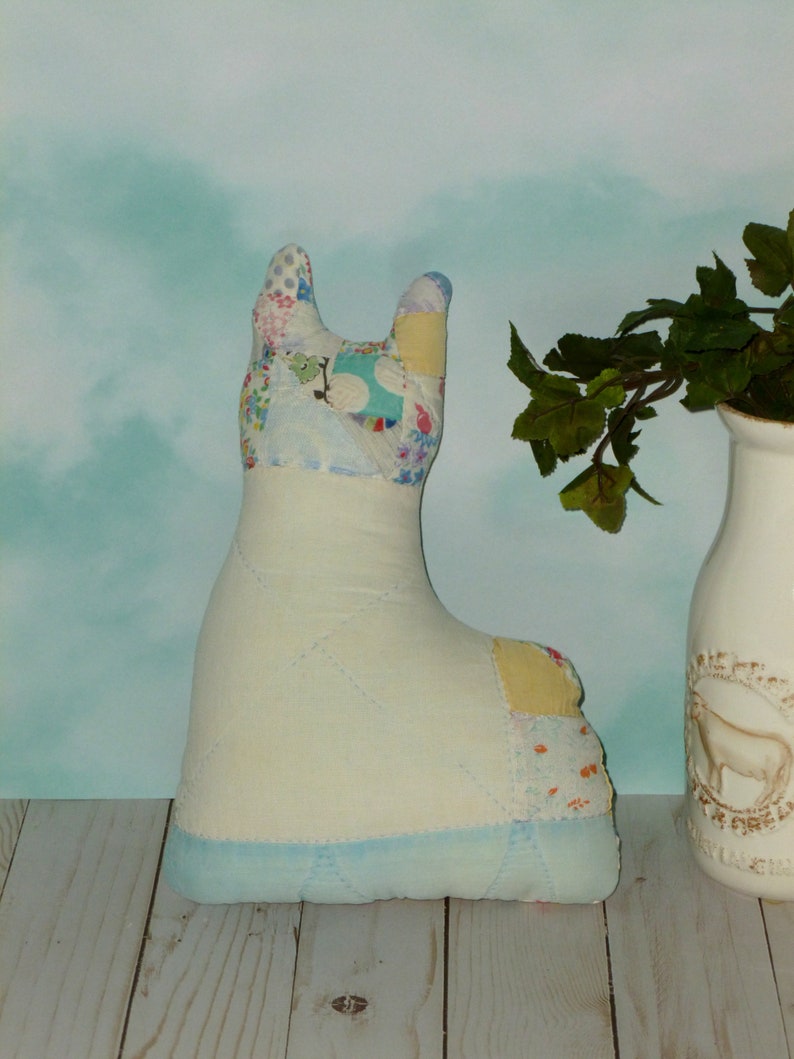 May include: A handmade patchwork fabric toy in the shape of a llama. The llama is light blue with yellow, pink, and white patchwork fabric. The llama is sitting on a light wood surface.