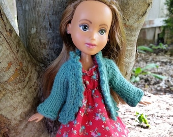 May include: A doll wearing a teal knitted cardigan over a red floral dress and pink shoes. The doll is standing near a tree trunk.