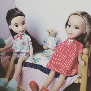 Puede incluir: Dos muñecas con pijamas están sentadas en una cama con un oso de peluche. Una muñeca lleva un pijama floral blanco y rosa y la otra muñeca lleva un pijama de lunares rojos y blancos.