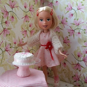 May include: A doll with blonde hair and a pink and white dress stands in front of a pink and white cake on a pink and white table. The doll is wearing a white cardigan and has red hair clips in her hair.