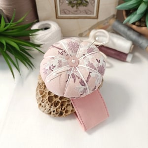 Può includere: Un cuscino per spilli floreale rosa con un bottone al centro e una base in tessuto rosa. Il cuscino per spilli è appoggiato su una superficie beige testurizzata.