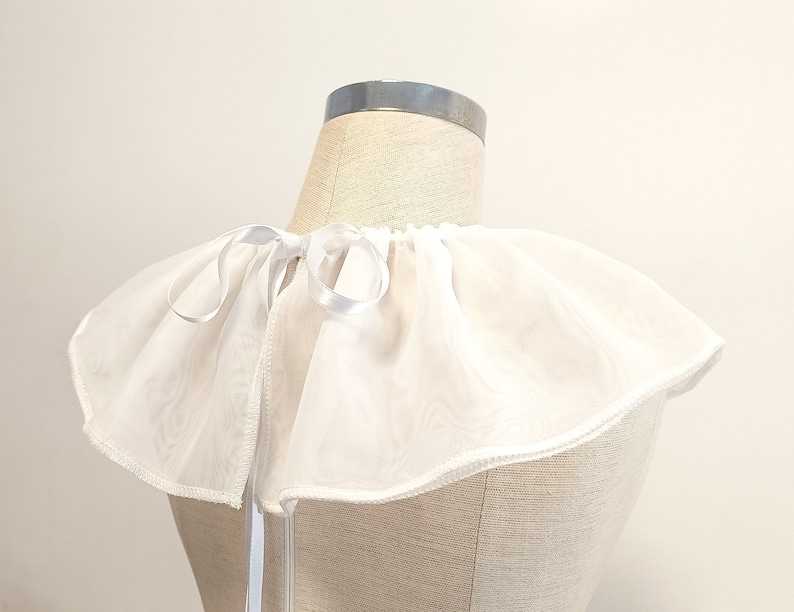 May include: White ruffled collar with a white satin ribbon tie. The collar is made of a sheer fabric and has a delicate, feminine look.