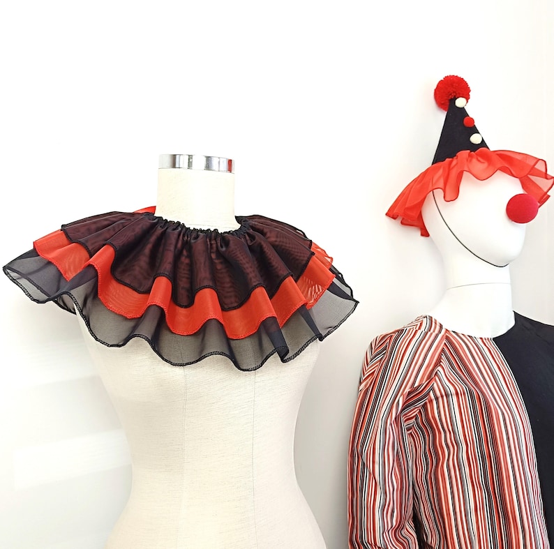 May include: A black and red ruffled collar with a layered design. The collar is on a white mannequin. A clown costume with a red and white hat is in the background.