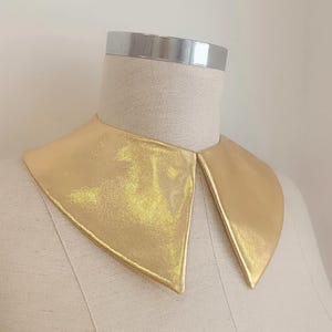 May include: A gold-colored, metallic-looking collar with a pointed design. The collar is displayed on a mannequin with a silver band around the neck. The collar has a shiny, textured appearance.