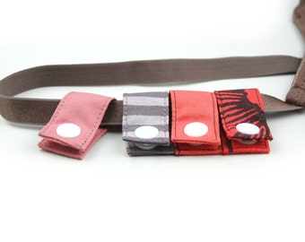 May include: Four fabric straps with white buttons, in pink, gray, red, and red with a black pattern. The straps are attached to a brown elastic band.