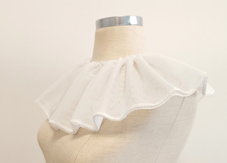 May include: White ruffled collar made of sheer fabric. The collar is sewn with a scalloped edge.