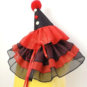 May include: A red and black layered ruffle collar with a black cone hat with a red pom-pom on top. The hat has white and red polka dots.