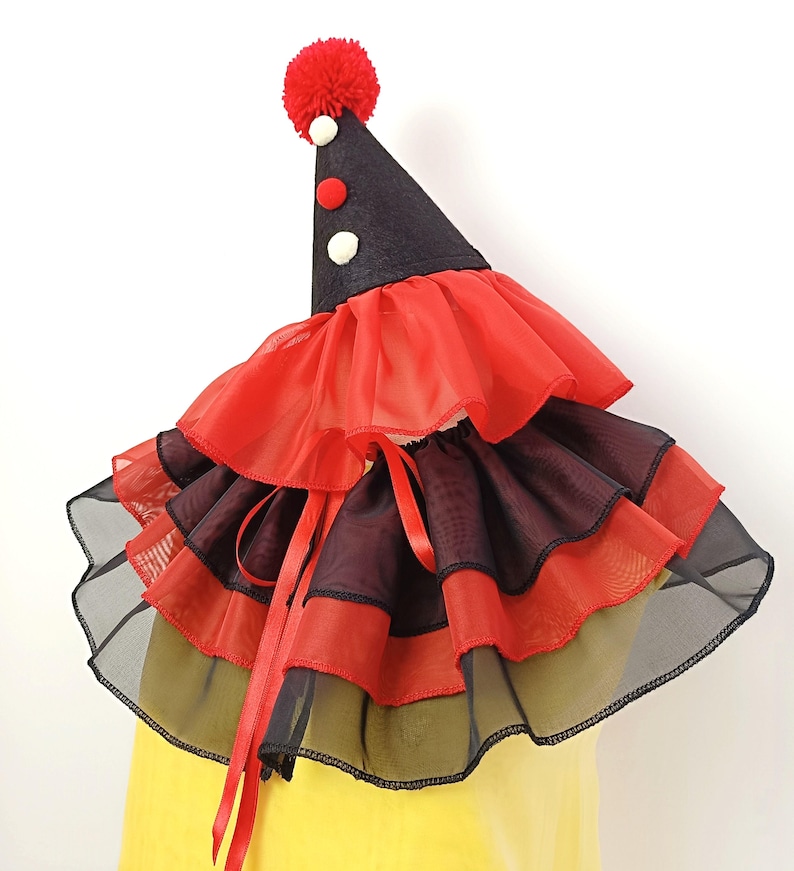 May include: A red and black layered ruffle collar with a black cone hat with a red pom-pom on top. The hat has white and red polka dots.