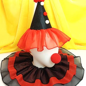 May include: A black and red clown hat with a red pom-pom on top. The hat has a red and black ruffle around the bottom. A red foam clown nose is attached to a black and red ruffle collar.