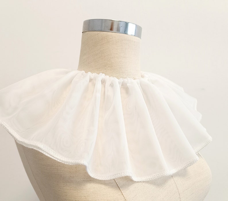 May include: A white ruffled collar made of sheer fabric. The collar is on a dress form and has a scalloped edge.