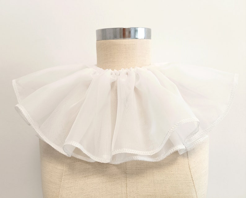 May include: A white ruffled collar made of sheer fabric. The collar is on a dress form.