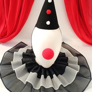 May include: A black and white clown collar with red pom poms and a red nose. The collar is made of tulle and has multiple layers. The collar is on a white mannequin head with a black and red cone hat.