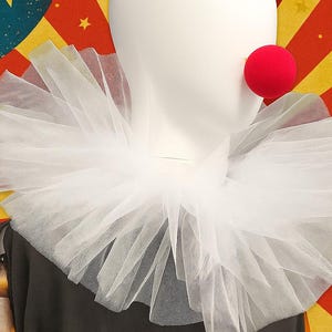 Könnte beinhalten: Ein weißer Clownkragen aus Tüll und eine rote Nase auf einem Schaufensterkopf. Der Hintergrund ist ein Zirkus-Thema-Design mit roten, gelben und blauen Farben und Sternen. Der Kragen ist geschichtet und voluminös.