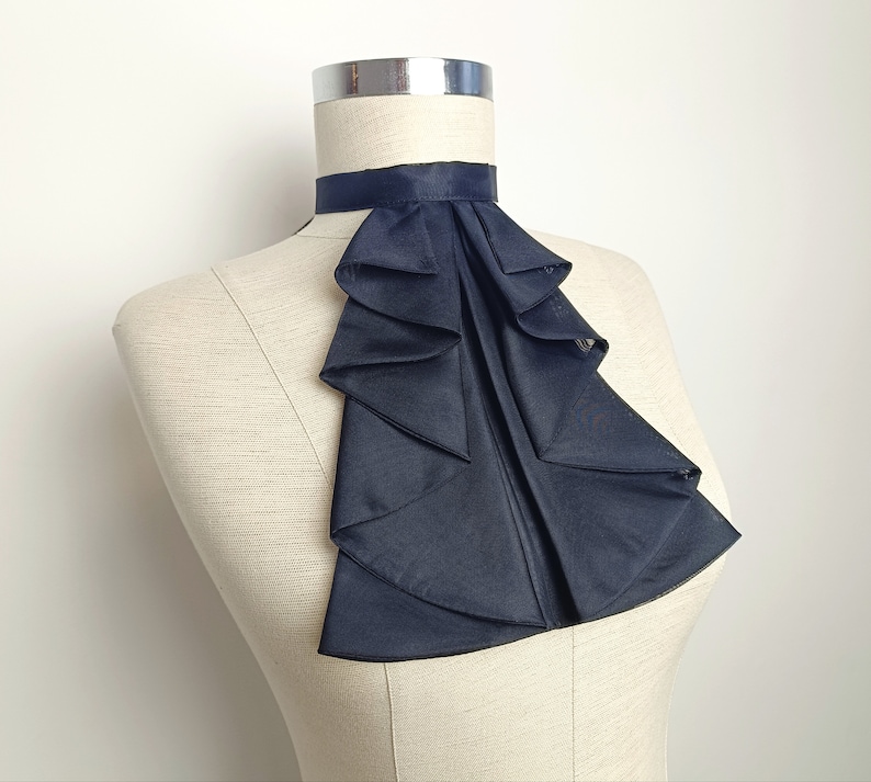 May include: A black ruffled fabric collar, also known as a cravat, is displayed on a dress form. The collar is made of a soft, flowing fabric and has a layered, cascading design.