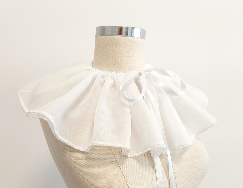 May include: A white ruffled collar with a white satin ribbon tie. The collar is made of a soft, sheer fabric and is perfect for adding a touch of elegance to any outfit.