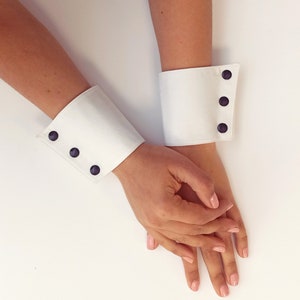 Puede incluir: Dos puños de camisa de tela blanca con botones negros. Los puños se llevan en las muñecas de una persona.