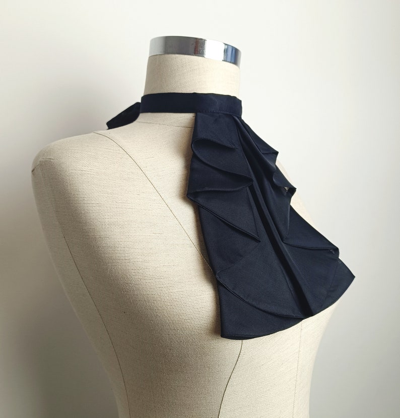 May include: A black ruffled collar, worn on a white mannequin. The collar is made of a soft, flowing fabric and has a high, dramatic neckline.