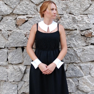 Puede incluir: Un vestido negro con un corpiño texturizado y una falda fluida. El vestido se complementa con un cuello blanco y puños blancos a juego. La mujer está de pie frente a una pared de piedra.