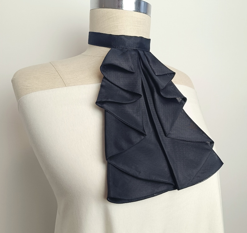 May include: A black ruffled fabric collar, worn as a neck accessory. The collar is made of multiple layers of fabric, creating a dramatic and elegant look.