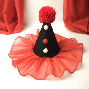 May include: A black felt cone hat with a red pom-pom on top. The hat has three white and red pom-poms on the front. The hat is sitting on a red ruffled fabric.