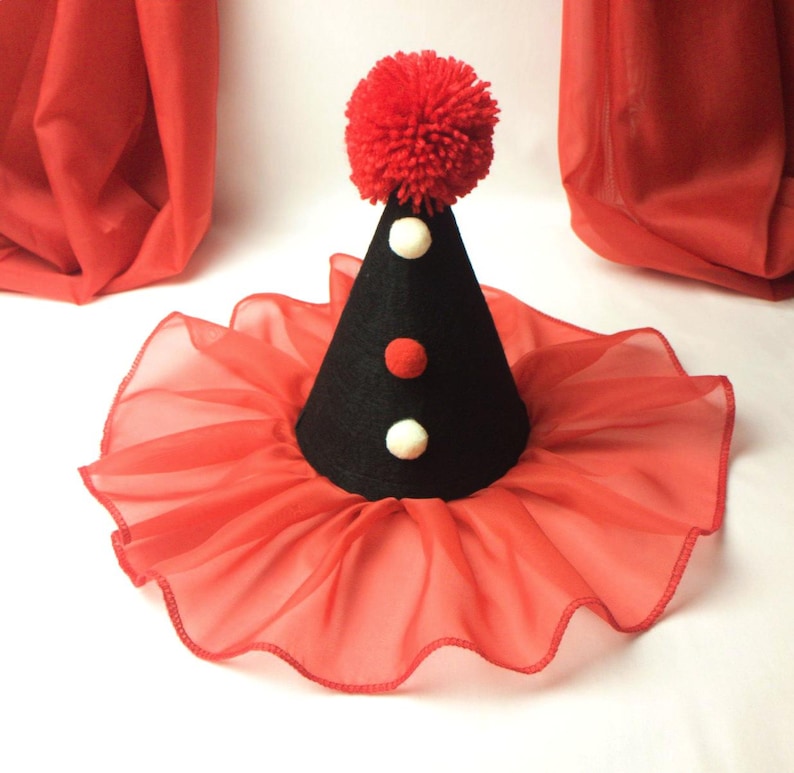 May include: A black felt cone hat with a red pom-pom on top. The hat has three white and red pom-poms on the front. The hat is sitting on a red ruffled fabric.
