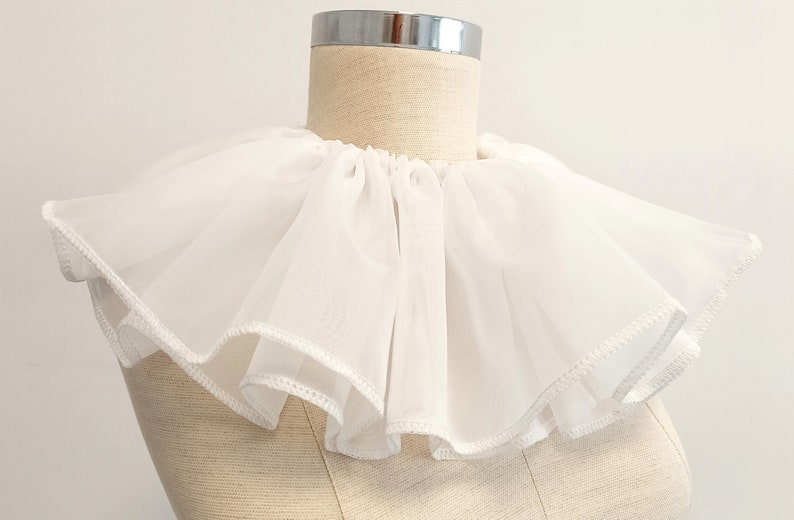 May include: A white ruffled collar made of sheer fabric. The collar is gathered at the neck and has a scalloped edge.