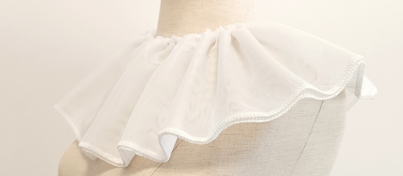 May include: White ruffled collar made of sheer fabric. The collar has a scalloped edge and is sewn with white thread.