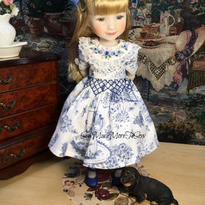 May include: A porcelain doll wearing a blue and white dress with a lace collar and blue bows in her hair. The doll is standing on a floral patterned rug with a small black dachshund figurine beside her.