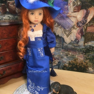 May include: A porcelain doll wearing a blue dress with white embroidery and a blue hat with feathers. The doll has red hair and blue eyes. A small black dog figurine is sitting on a white and blue patterned surface.