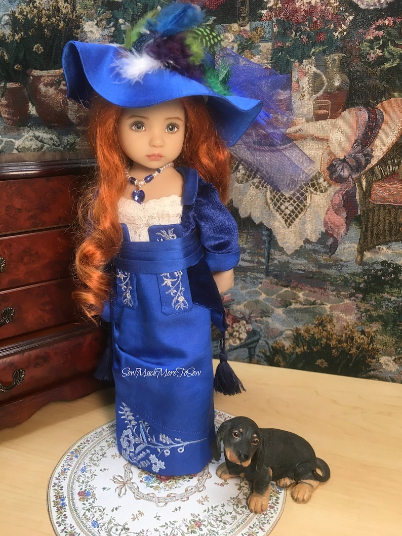 May include: A porcelain doll wearing a blue dress with white embroidery and a blue hat with feathers. The doll has red hair and blue eyes. A small black dog figurine is sitting on a white and blue patterned surface.